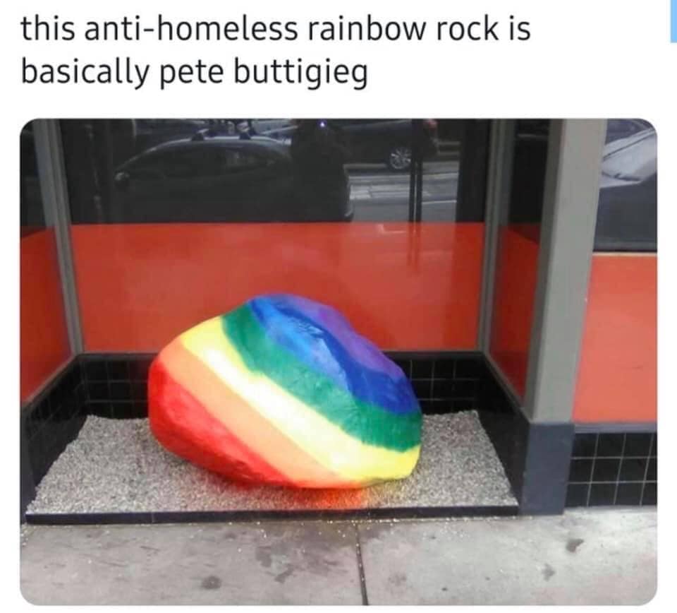 The meme is a photo of a large, grey rock with a rainbow-colored paint job, resembling a giant rainbow rock. The image is accompanied by the text: "This anti-homeless rainbow rock is basically Pete Buttigieg." The joke is that the rock, which is often used as a decorative feature in public spaces to promote a sense of community and inclusivity, is being sarcastically referred to as "basically" Pete Buttigieg, a politician known for his progressive policies.